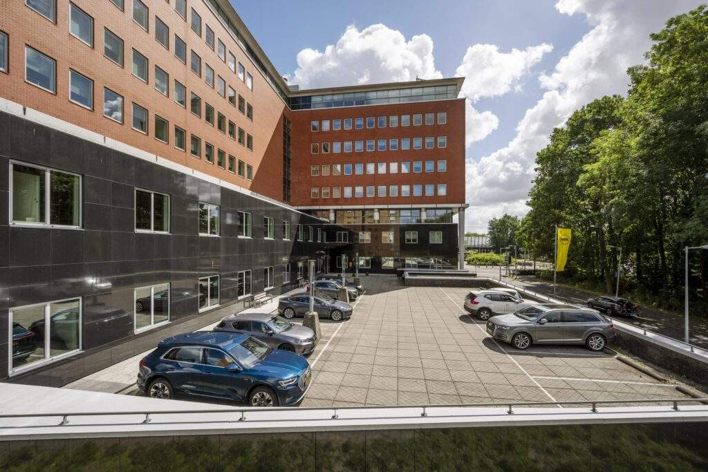Parkeerterrein met geparkeerde auto's naast een modern kantoorgebouw aan het Koningin Wilhelminaplein.